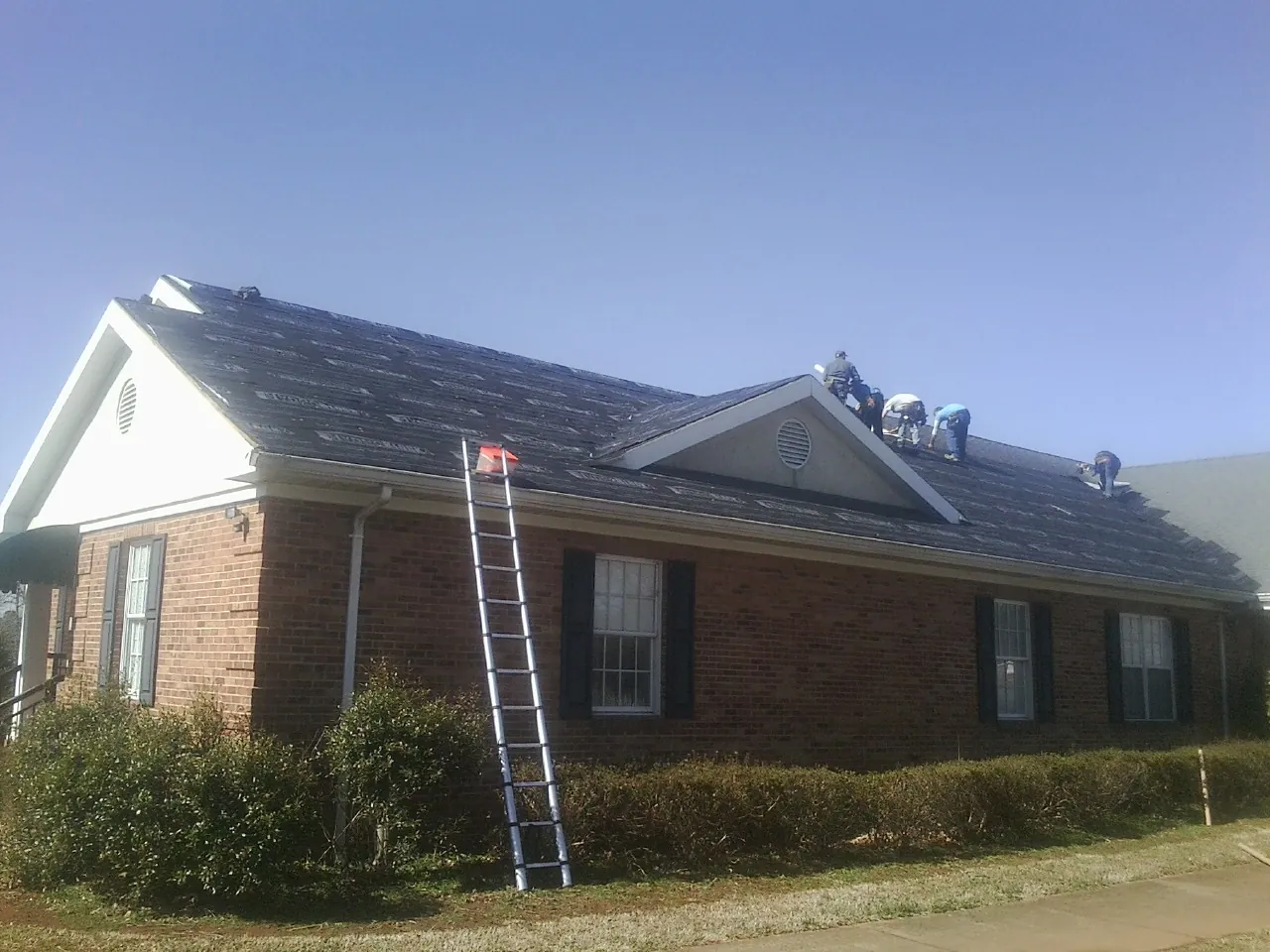Crew tearing shingles off a brick ranch roof with a ladder set — Emergency Roof Repair work by our crew in Mc Bain