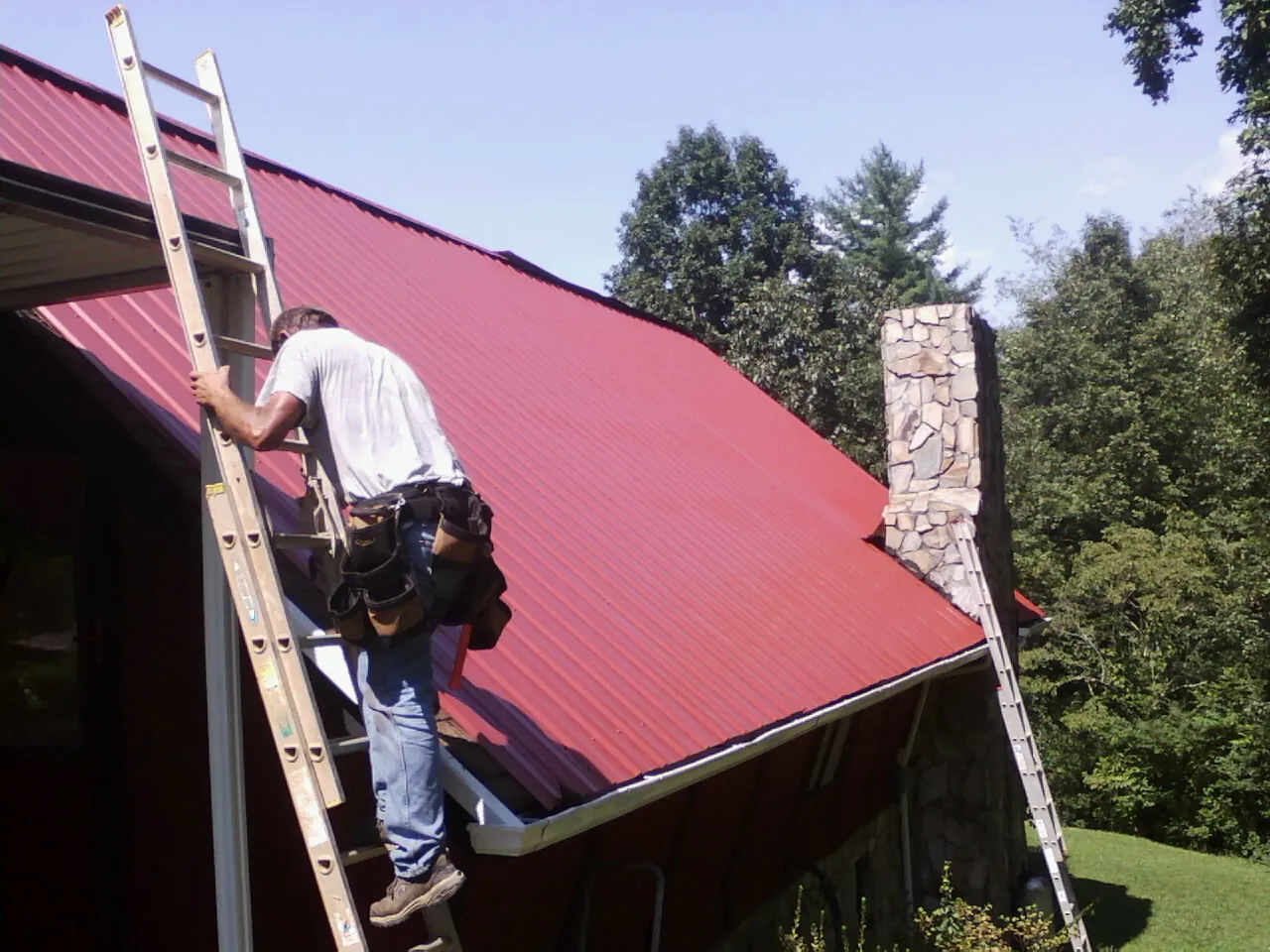 Roofer on a ladder beside a red metal roof and stone chimney — Soffit & Fascia Repair by Roofing Brothers in Mc Bain